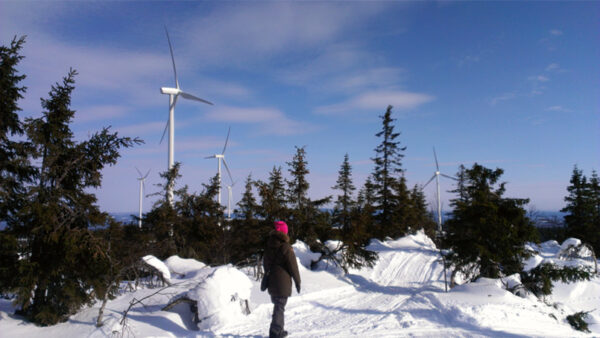 Snöigt landskap med en person som promenerar, vindkraftverk i bakgrunden.